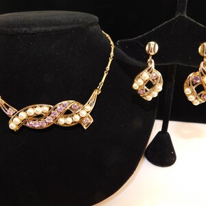 May include: A gold tone necklace and earrings set with a twisted design featuring faux pearls and purple rhinestones.