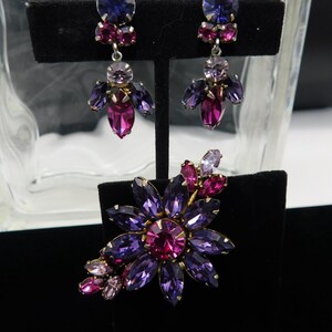Op de afbeelding: Een set vintage oorbellen en een broche met een bloemmotief met paarse, roze en heldere strass.