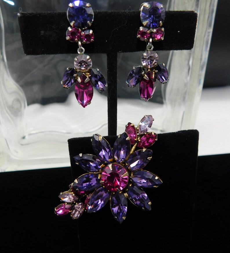 Op de afbeelding: Een set vintage oorbellen en een broche met een bloemmotief met paarse, roze en heldere strass.