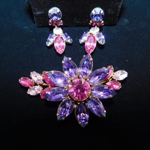Op de afbeelding: Een paarse en roze bloemvormige broche met strass. De broche is gemaakt van metaal en heeft een gouden afwerking.