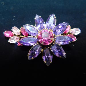 Op de afbeelding: Een paarse en roze broche in de vorm van een bloem met strass. De broche heeft een gouden setting.