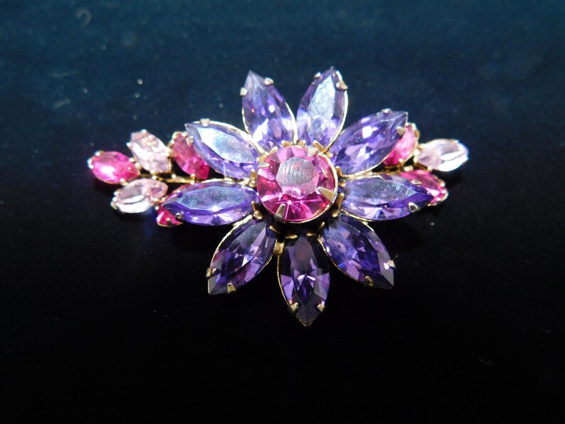 Op de afbeelding: Een paarse en roze broche in de vorm van een bloem met strass. De broche heeft een gouden setting.