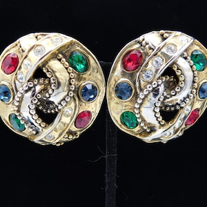 May include: A pair of gold-tone clip-on earrings with a multi-colored stone design. The earrings feature red, green, and blue stones set in a gold-tone metal base.