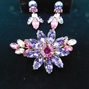 Op de afbeelding: Een broche in de vorm van een paarse en roze bloem met een grote centrale roze strasssteen omgeven door kleinere paarse strassstenen. De broche is versierd met kleinere roze strassstenen aan de randen. De broche wordt geleverd met een paar oorbellen met een vergelijkbaar ontwerp.
