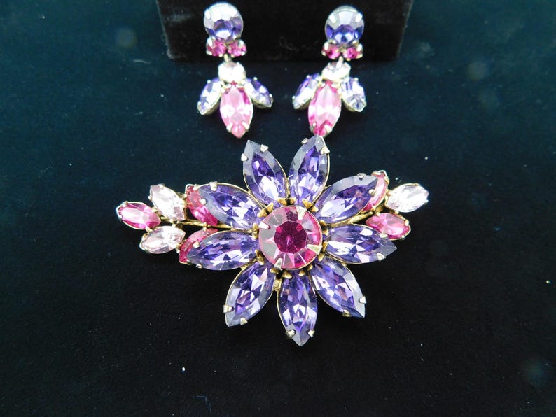 Op de afbeelding: Een broche in de vorm van een paarse en roze bloem met een grote centrale roze strasssteen omgeven door kleinere paarse strassstenen. De broche is versierd met kleinere roze strassstenen aan de randen. De broche wordt geleverd met een paar oorbellen met een vergelijkbaar ontwerp.