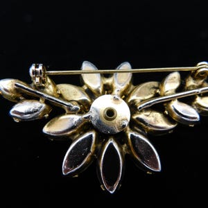 Op de afbeelding: Een broche van goudkleurige metaal met een cluster van heldere en zwarte strass in de vorm van een bloem. De broche heeft een pin en sluiting aan de achterkant.