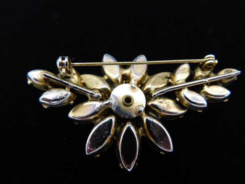 Op de afbeelding: Een broche van goudkleurige metaal met een cluster van heldere en zwarte strass in de vorm van een bloem. De broche heeft een pin en sluiting aan de achterkant.