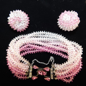 May include: A vintage jewelry set featuring a bracelet and matching earrings. The bracelet is composed of multiple strands of clear and pink faceted beads, with a gradient effect. The earrings are round, also made of faceted beads. The set has a silver clasp.