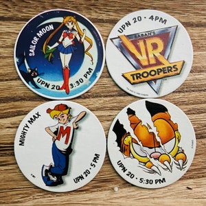 May include: Four circular collectible tokens featuring cartoon characters and logos. One token shows Sailor Moon, another VR Troopers, Mighty Max, and a tiger claw. Each token has the UPN 20 logo and a time.