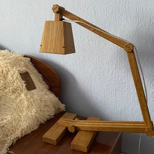 Op de afbeelding: Een houten bureaulamp met een rechthoekige kap en een flexibele arm. De lamp is gemaakt van natuurlijk hout en heeft een wit snoer. De lamp staat op een houten tafel.