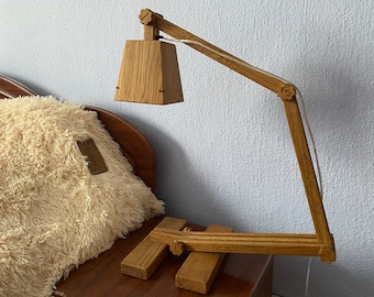 May include: A wooden desk lamp with a rectangular shade and a flexible arm. The lamp is made of natural wood and has a white cord. The lamp is sitting on a wooden table.