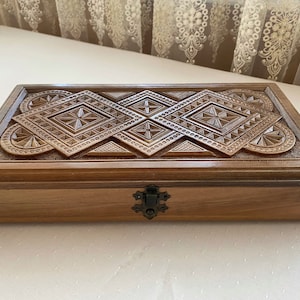 Puede incluir: Una caja de madera con intrincados tallados en la tapa. La caja tiene un acabado marrón oscuro y un cierre de metal.