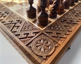 May include: A wooden chessboard with intricate carvings on the edges. The board is made of dark wood and has a light wood inlay. The chess pieces are also made of wood and are arranged on the board.
