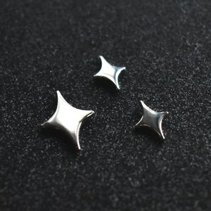 May include: Three silver-toned metal star-shaped embellishments with four pointed tips. The stars vary in size, featuring a smooth, reflective surface that catches the light. These small, decorative pieces have a distinct four-pointed sparkle design.