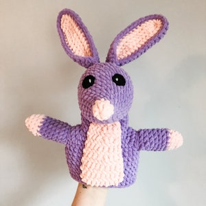 May include: Purple and pink crocheted bunny puppet with black eyes. The puppet is being held up by a hand.