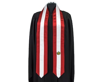 May include: A graduation sash with red, white, and red vertical stripes, draped over a black academic gown. The sash features a small emblem with a crest and the colors of the Peruvian flag.