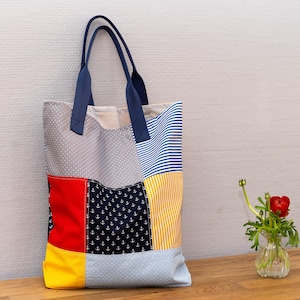 Könnte beinhalten: Eine Patchwork-Tragetasche aus blauem, gelbem, rotem und grauem Stoff. Die Tasche hat einen marineblauen Henkel und besteht aus verschiedenen gemusterten Stoffen.