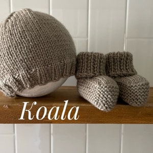 May include: A knitted baby set in a neutral beige color, including a beanie hat and a pair of booties. The word "Koala" is written in white script on the wooden shelf. The set is made of a soft, textured yarn.