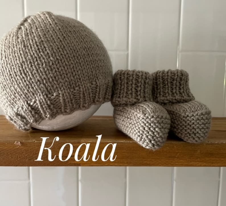 May include: A knitted baby set in a neutral beige color, including a beanie hat and a pair of booties. The word "Koala" is written in white script on the wooden shelf. The set is made of a soft, textured yarn.