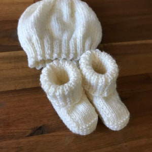 May include: A knitted baby set in cream color, including a beanie hat and a pair of booties. The hat has a ribbed brim and a rounded crown. The booties have a folded-over cuff. The set is made of soft yarn.
