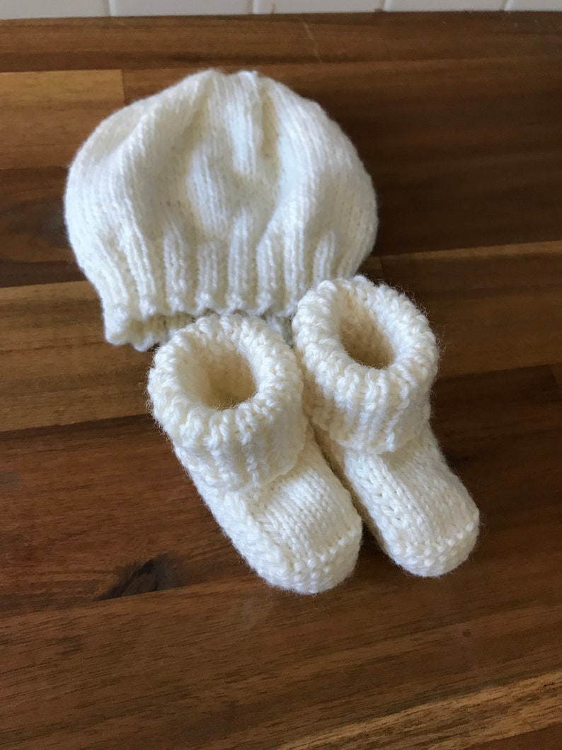 May include: A knitted baby set in cream color, including a beanie hat and a pair of booties. The hat has a ribbed brim and a rounded crown. The booties have a folded-over cuff. The set is made of soft yarn.