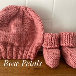 May include: A knitted baby set in a rose pink color. The set includes a beanie-style hat and a pair of booties. The text "Rose Petals" is displayed on the bottom left of the image. The items are made of a soft, textured yarn.