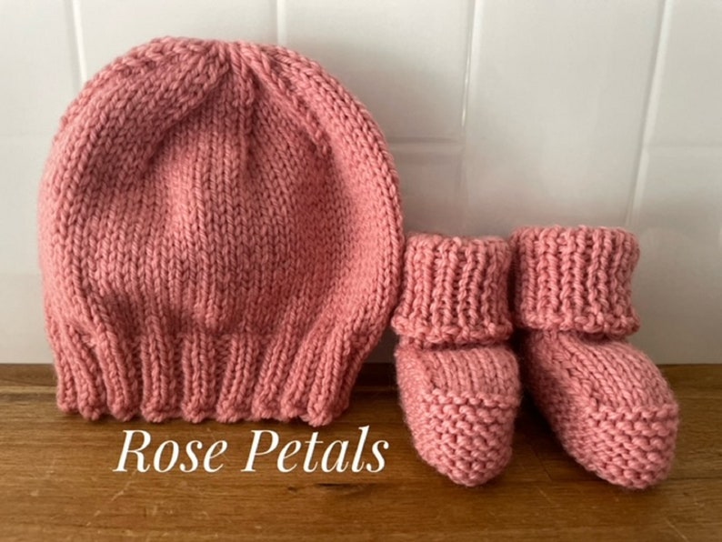 May include: A knitted baby set in a rose pink color. The set includes a beanie-style hat and a pair of booties. The text "Rose Petals" is displayed on the bottom left of the image. The items are made of a soft, textured yarn.