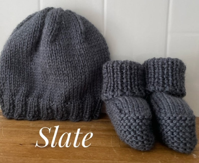 May include: A knitted slate gray baby hat and matching booties. The hat is a simple beanie style, and the booties have a folded cuff. The word "Slate" is written in cursive at the bottom of the image.