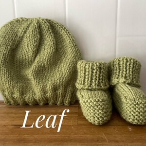 May include: A knitted baby set in a soft green color, including a beanie hat and a pair of booties. The word "Leaf" is written in white script at the bottom. The set is made of a chunky knit material.
