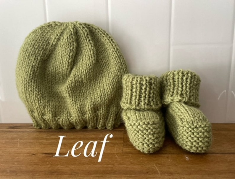 May include: A knitted baby set in a soft green color, including a beanie hat and a pair of booties. The word "Leaf" is written in white script at the bottom. The set is made of a chunky knit material.