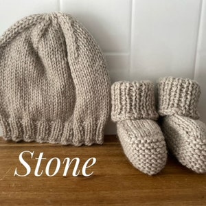 May include: A knitted baby hat and booties set in a neutral stone color. The hat is a beanie style with a ribbed brim. The booties have a folded-over cuff. The set is made of a soft, textured yarn.