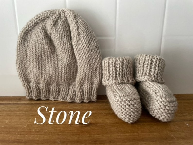 May include: A knitted baby hat and booties set in a neutral stone color. The hat is a beanie style with a ribbed brim. The booties have a folded-over cuff. The set is made of a soft, textured yarn.