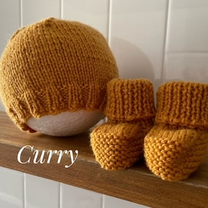 May include: Mustard yellow knitted baby hat and booties set. The hat is round and the booties have a folded cuff. The word "Curry" is written in cursive on the wooden shelf. The set is made of a soft, textured yarn.