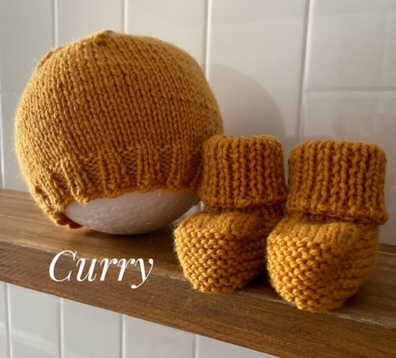 May include: Mustard yellow knitted baby hat and booties set. The hat is round and the booties have a folded cuff. The word "Curry" is written in cursive on the wooden shelf. The set is made of a soft, textured yarn.