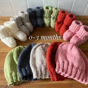 May include: A collection of knitted baby booties and beanies in various colors, including cream, gray, navy, green, red, and pink. The text "0-3 months" is displayed. These items are likely for newborns or infants.