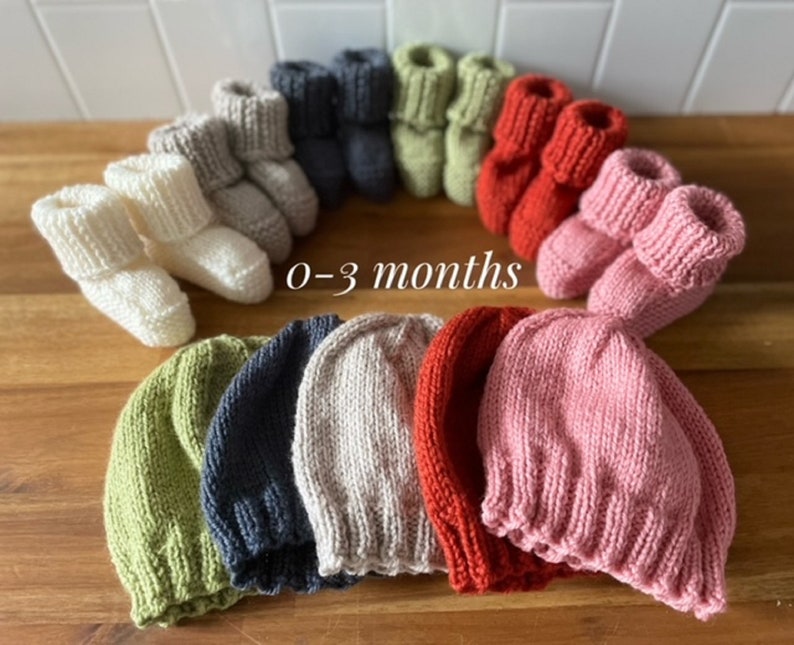 May include: A collection of knitted baby booties and beanies in various colors, including cream, gray, navy, green, red, and pink. The text "0-3 months" is displayed. These items are likely for newborns or infants.