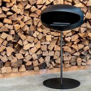 May include: A black, round, free-standing fireplace with a modern design. The fireplace is on a stand with a round base. The fireplace is in front of a wall of stacked firewood.