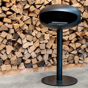 May include: A black metal fire pit with a round, bowl-shaped top and a tall, cylindrical stand. The fire pit is sitting on a concrete surface in front of a wall of stacked firewood.