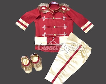 May include: A red and gold baby outfit with a matching pair of shoes. The outfit features a red shirt with gold trim and a gold belt with a letter 'A' on it. The trousers are gold with red stripes. The shoes are gold with brown fur trim.