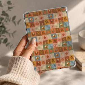 May include: A clear tablet case featuring a repeating patchwork pattern of autumn-themed icons including orange pumpkins, slices of pumpkin pie, brown candles, red mushrooms, yellow flowers, and fall leaves against a grid of blue and beige squares.