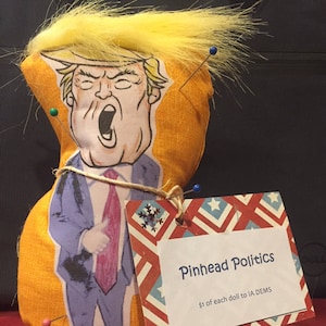 May include: A yellow fabric doll with a cartoon image of a man with a red tie and a yellow wig. The doll is pinned with colorful pins. A sign reads "Pinhead Politics $1 of each doll to IA DEMS".