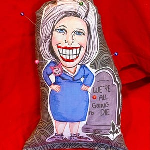 May include: A fabric doll with a cartoon illustration of a woman in a blue dress, with a large smile and white hair. The doll has pins in it and a tombstone that reads "WE'RE ALL GOING TO DIE".