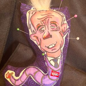 Puede incluir: Muñeca de tela hecha a mano con una ilustración de dibujos animados de un hombre con pelo blanco y un traje. La muñeca es morada con un patrón de remolino y las letras "RFK" son visibles. Varios alfileres están insertados en la muñeca.
