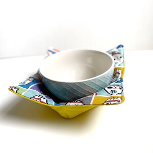May include: A yellow bowl cozy with a colorful Toy Story print featuring characters like Woody and Buzz Lightyear. The cozy is holding a white ceramic bowl.