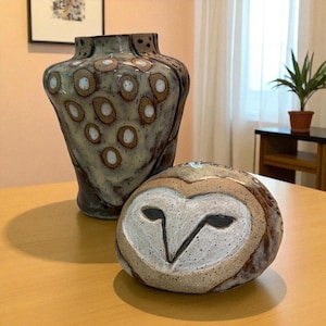 May include: A ceramic vase and owl sculpture. The vase is a mottled brown and tan with white circular accents. The owl sculpture has a white face with black eyes and a brown and tan body. Both are on a wooden table.