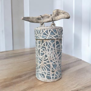 May include: A ceramic jar with a blue and white geometric pattern, with a lid and a piece of driftwood attached to the top with wire.