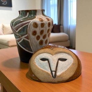 May include: A ceramic vase with a dark brown base and a patterned upper section in green, white, and maroon. Beside it is a ceramic owl sculpture with a white face and brown details. Both are on a wooden table.
