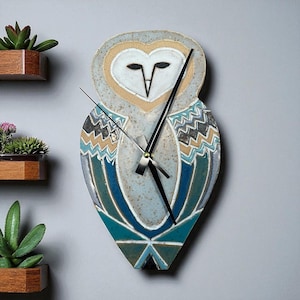 May include: A ceramic owl-shaped wall clock with a heart-shaped face and geometric wing patterns in blue, teal, tan, and grey. The clock has black hands and is mounted on a grey wall. Decorative wooden planters with succulents are visible.