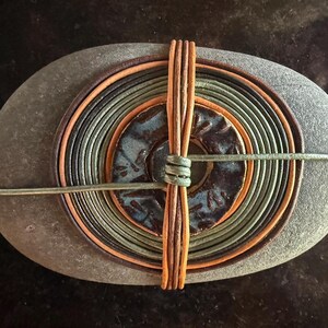 Peut inclure: Une pierre grise lisse de forme ovale ornée de cercles concentriques de fins cordons de cuir multicolores dans des tons marron, beige et vert olive. Une pièce centrale en céramique circulaire avec un vernis bleu foncé texturé est fixée.