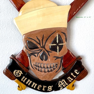 May include: A wooden wall plaque featuring a skull with a crosshair over one eye, a sailor's hat, and crossed wooden axes. The plaque is inscribed with the words "Gunners Mate" and "Officially Licensed by the U.S. Navy". It measures 15 inches by 12 inches.